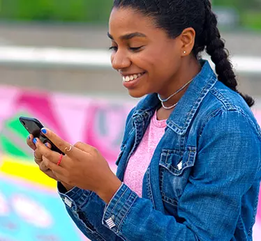 Young woman smiling at her smartphone wearing a pink shirt with a denim jacket.